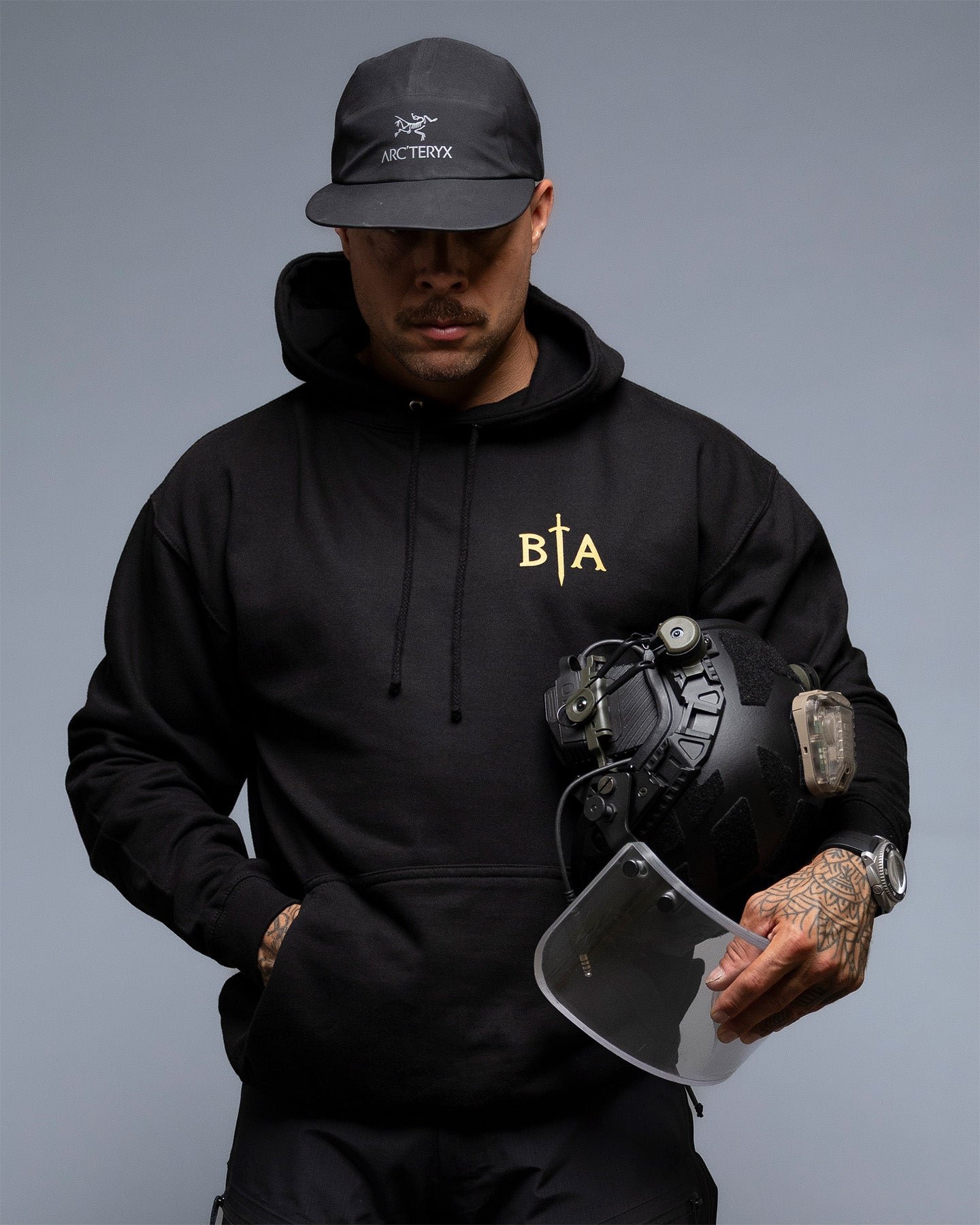 Person wearing a black hoodie with a logo, holding a helmet against a gray background. #color_black